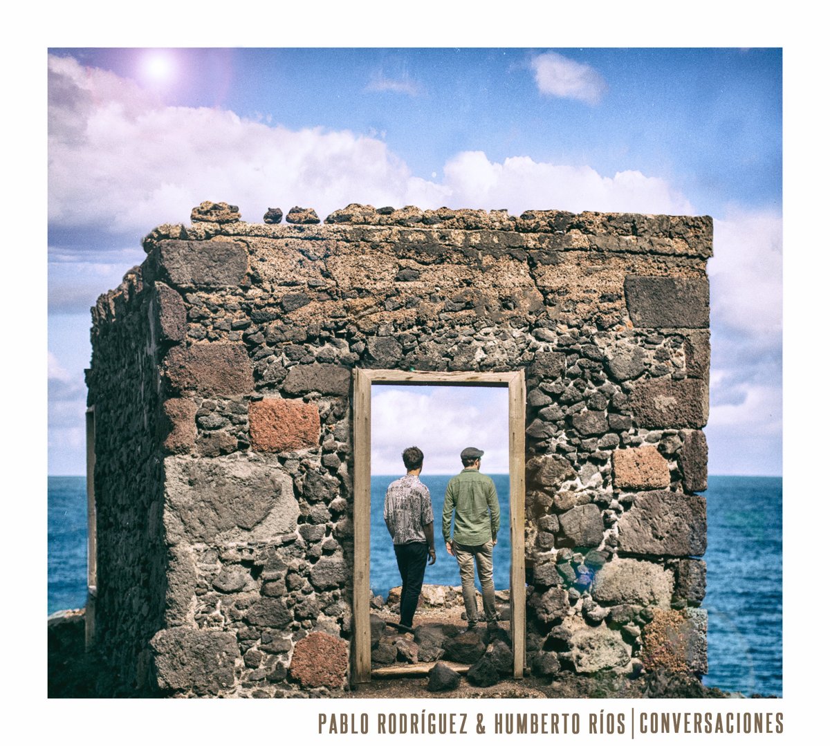 Conversaciones album cover: the duo standing in a stone doorway facing the ocean